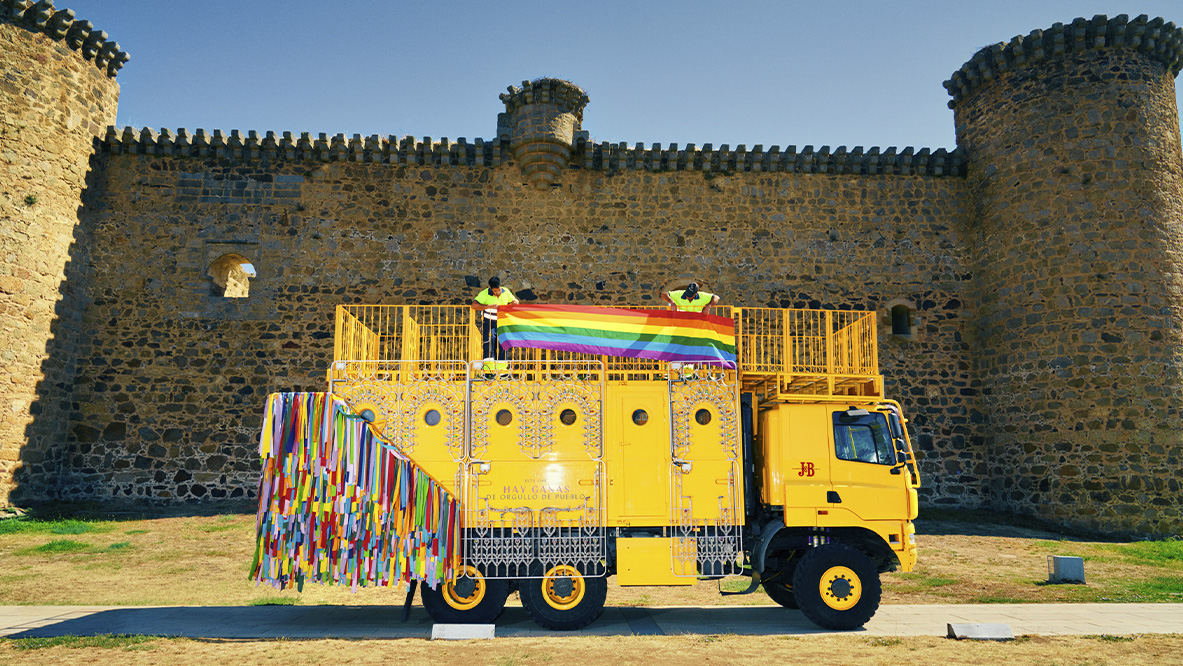 Dos operarios adornan la carroza de J&B con luces de colores y la bandera arcoíris. Están ante una enorme muralla antigua flanqueada por dos torreones circulares. En un lateral de la carroza pone 'Hay ganas de orgullo de pueblo',